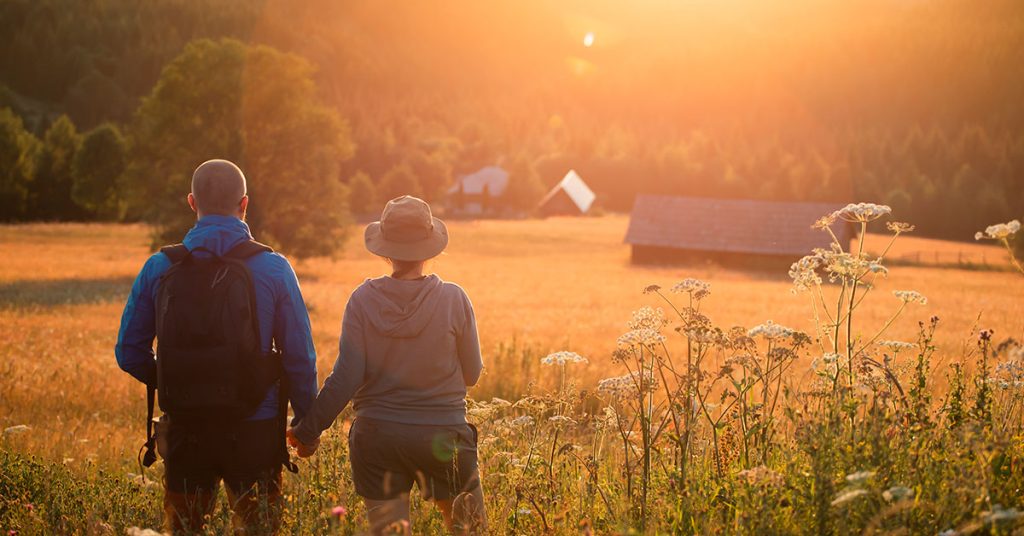Kaksi henkilöä kävelee käsi kädessä kukkaniityllä auringonnousun aikaan.